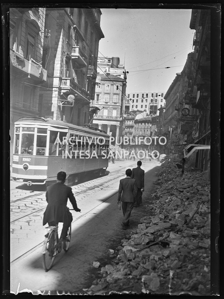 Scorcio di corso Vittorio Emanuele II, a Milano, all'indomani dei bombardamenti: le macerie degli edifici distrutti durante gli attacchi aerei sono state addossate ai lati della strada per consentire la circolazione, come in questa immagine, a tram e pedoni. Un tram tipo 1928, linea 3, avanza nella via