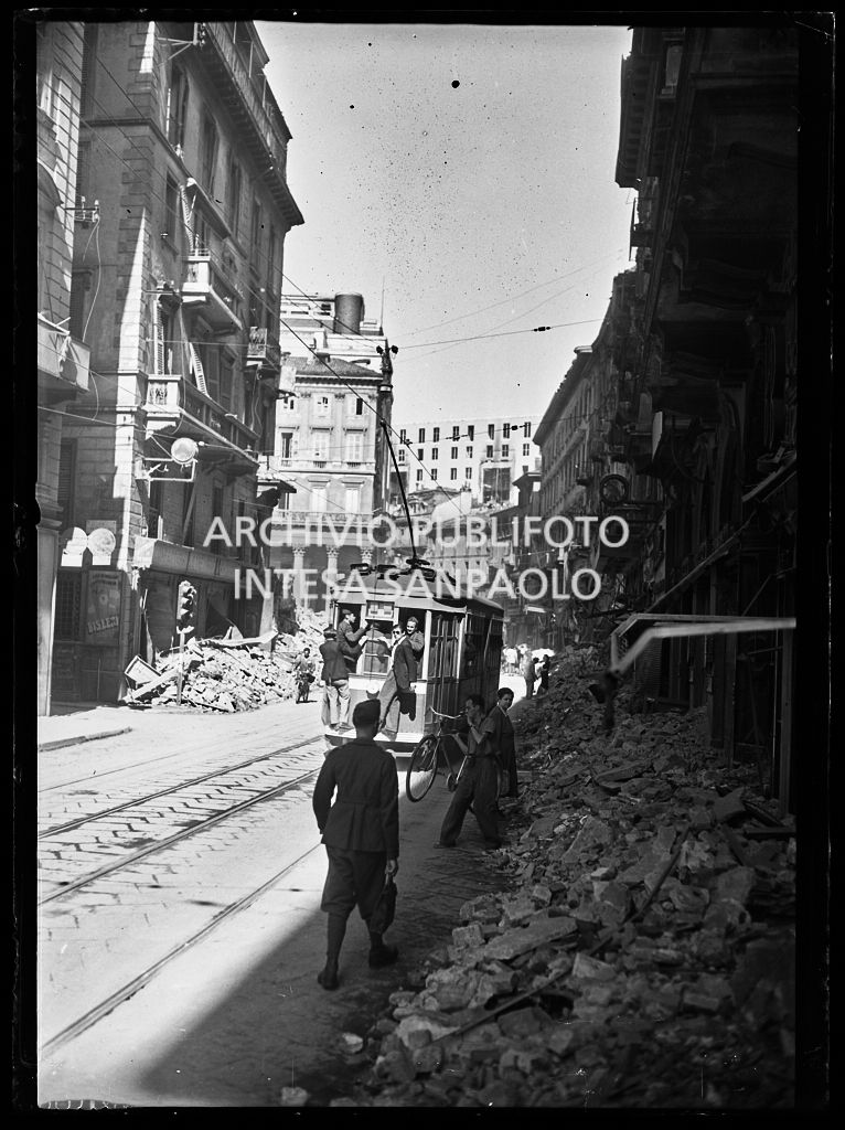 Scorcio di corso Vittorio Emanuele II, a Milano, all'indomani dei bombardamenti: le macerie degli edifici distrutti durante gli attacchi aerei sono state addossate ai lati della strada per consentire la circolazione, come in questa immagine, a tram, biciclette e pedoni. Un tram tipo 1928, linea 3, avanza nella via con passeggeri sul predellino posteriore della vettura