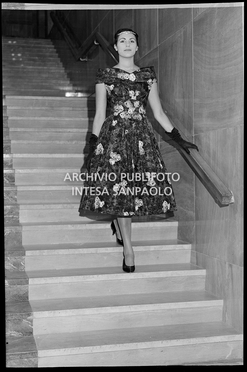 Indossatrice con abito fiorato, guanti e tiara mentre scende le scale<br>21791