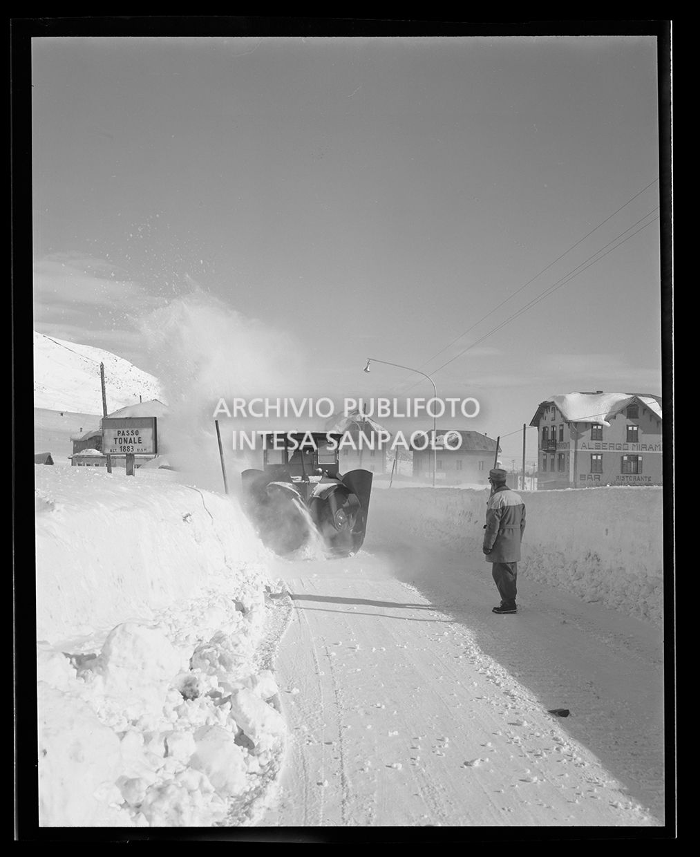 Spazzaneve in funzione su una strada mentre un uomo controlla il lavoro. Su un cartello si legge "A.N.A.S. Passo del Tonale alt. 1883 m.s.m.". Sulla destra in lontananza si intravede l'albergo Miramonti  (simile allo scatto 25247)<br>25240