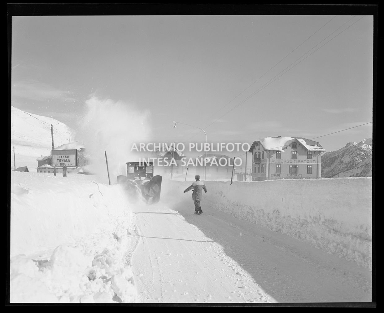 Spazzaneve in funzione su una strada mentre un uomo controlla il lavoro. Su un cartello si legge "A.N.A.S. Passo del Tonale alt. 1883 m.s.m.". Sulla destra si vede l'albergo Miramonti (simile allo scatto 25240)<br>25247