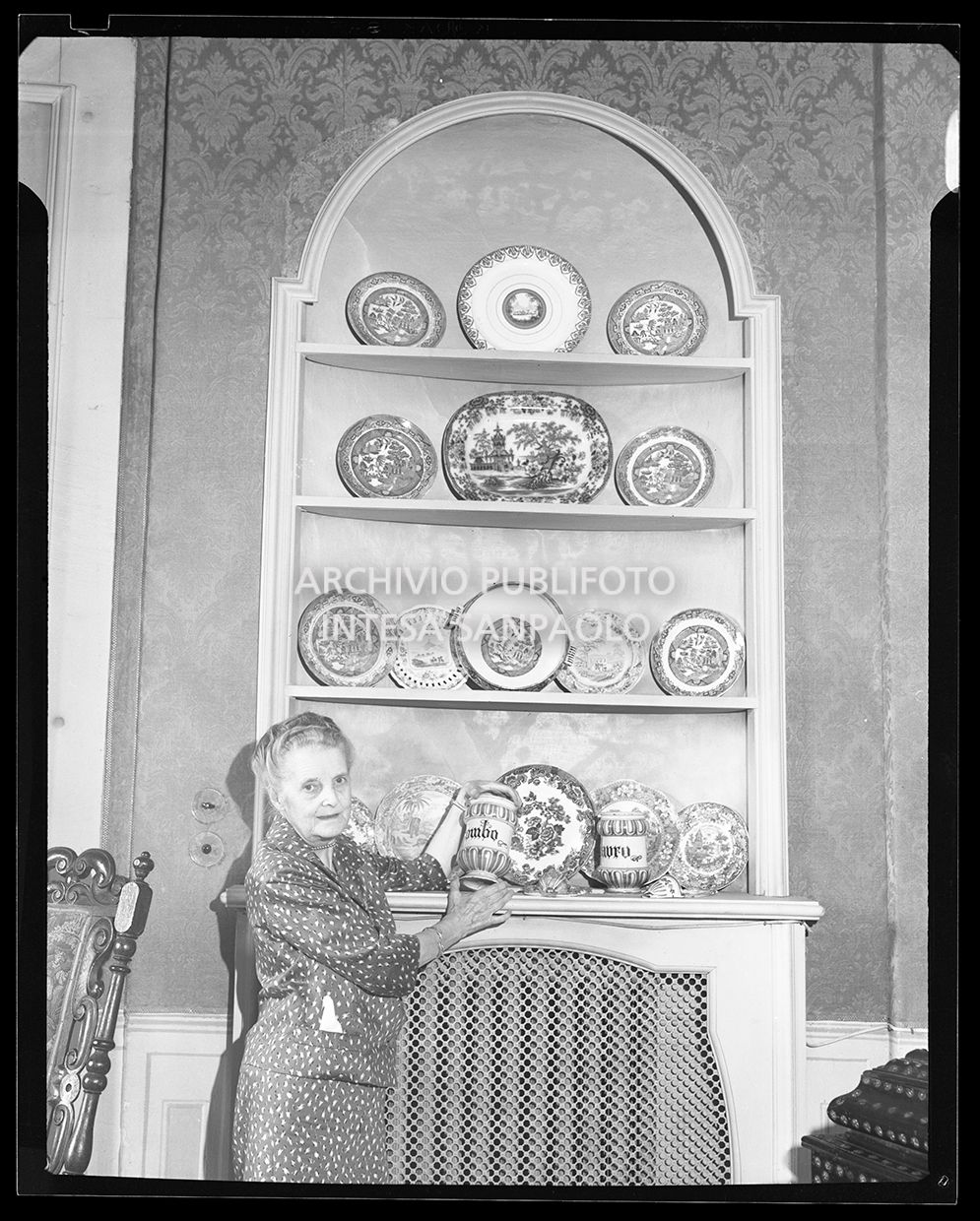 Signora mostra un contenitore in ceramica decorato di fronte ad una credenza piena di piatti decorati<br>28716