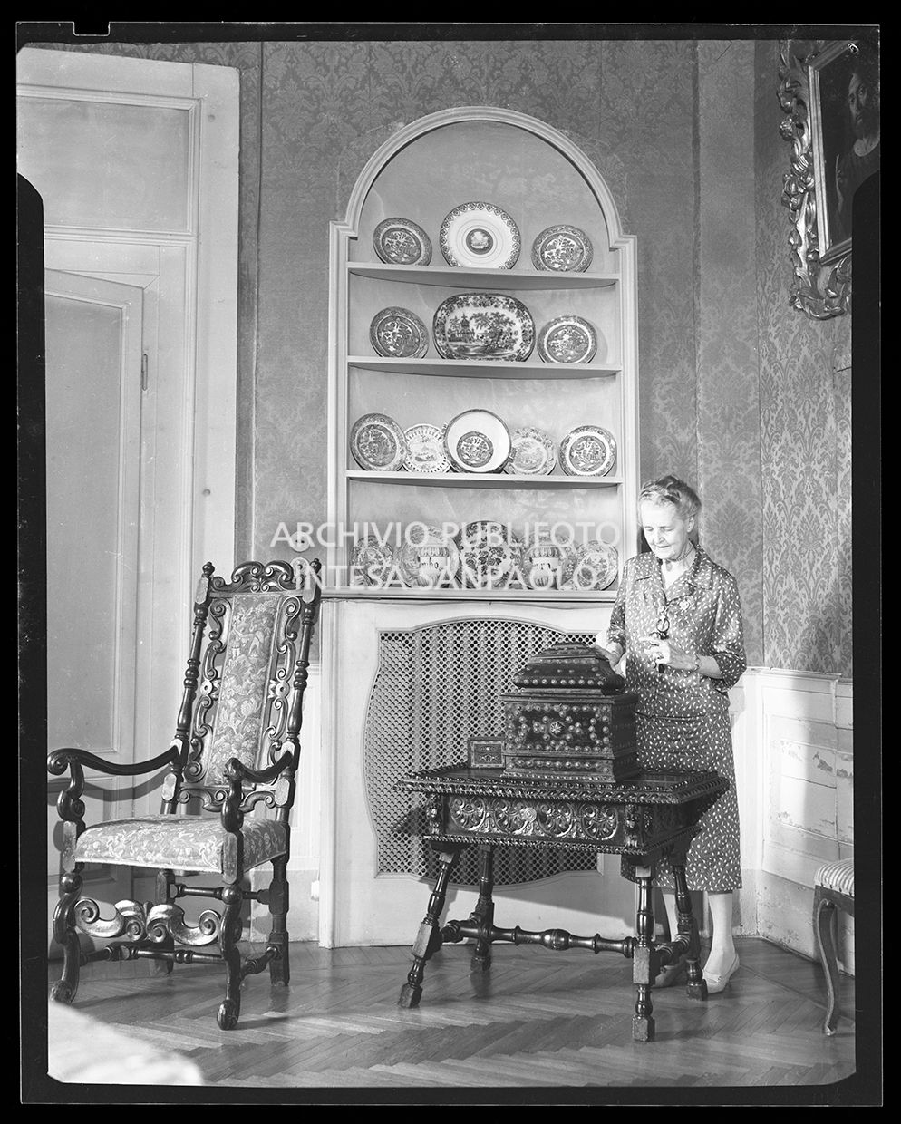 Signora mostra un soprammobile ed un tavolino di fronte ad una credenza piena di piatti decorati<br>28718