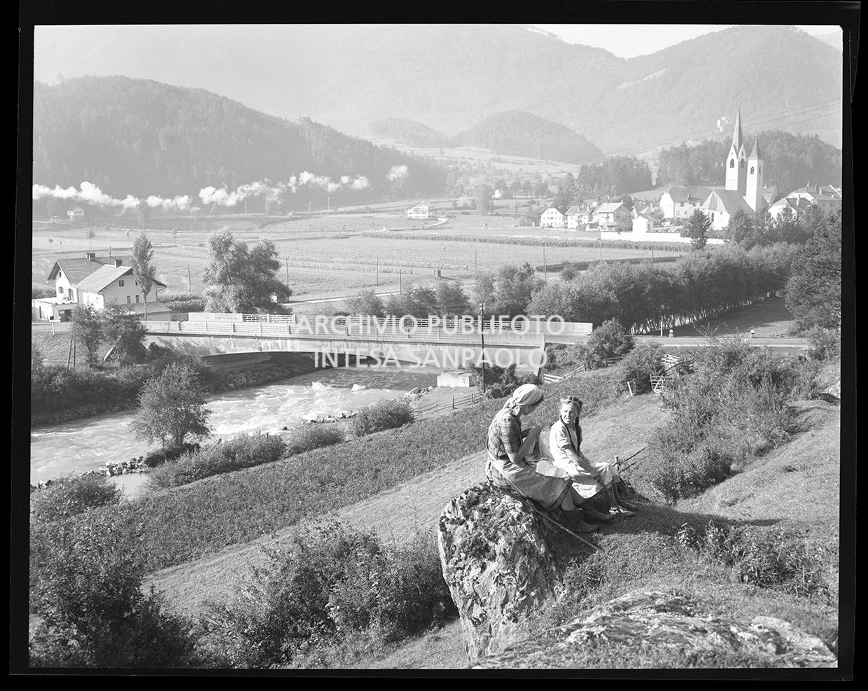 Una donna ed una bambina sedute su una roccia ed in lontananza un ponte e delle case<br>29867