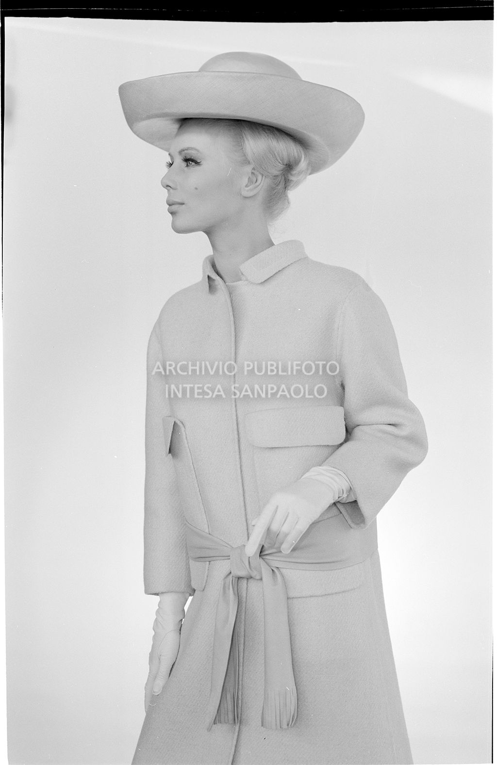 Indossatrice con cappotto legato sui fianchi, guanti e cappello<br>10913