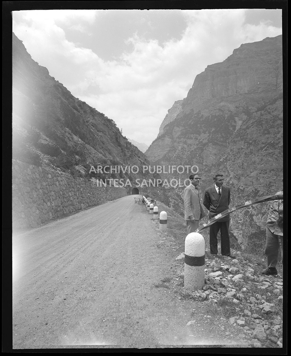 Sul ciglio della strada si vedono tre uomini ed in lontananza delle capre<br>19299