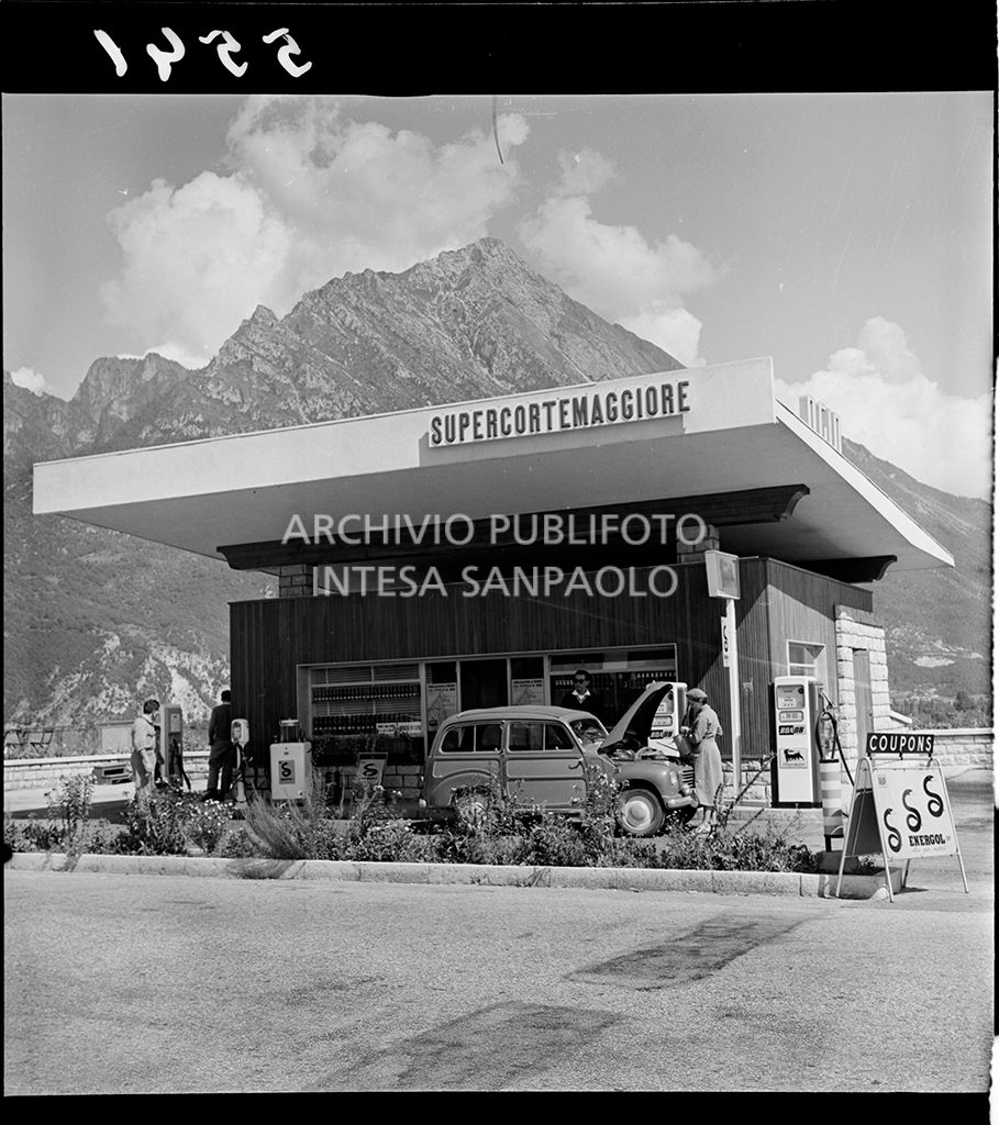 Automobile con cofano aperto ferma al distributore ed una donna con in mano un innaffiatoio<br>5541