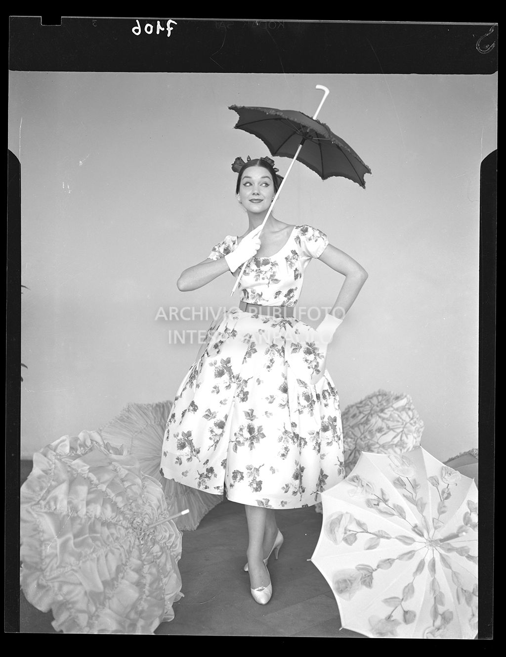 Donna con abito al polpaccio ampio e fiorato con guanti, cintura in vita e ombrello da sole. Altri ombrelli aperti ai suoi piedi<br>7106