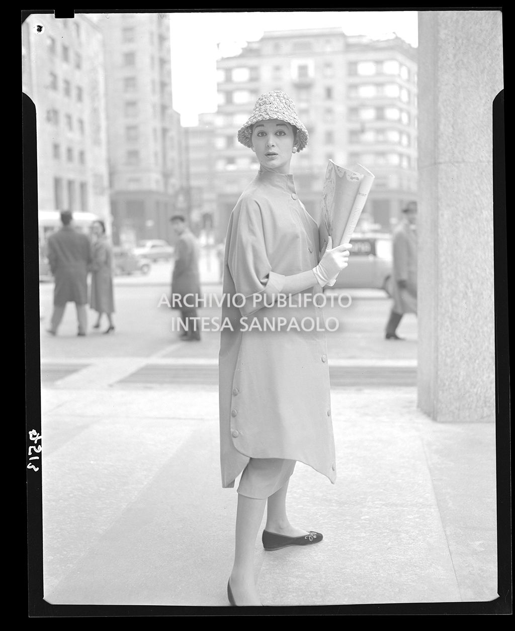 Donna con abito a camicia, cappello, guanti e ballerine; tiene in mano una rivista<br>7513