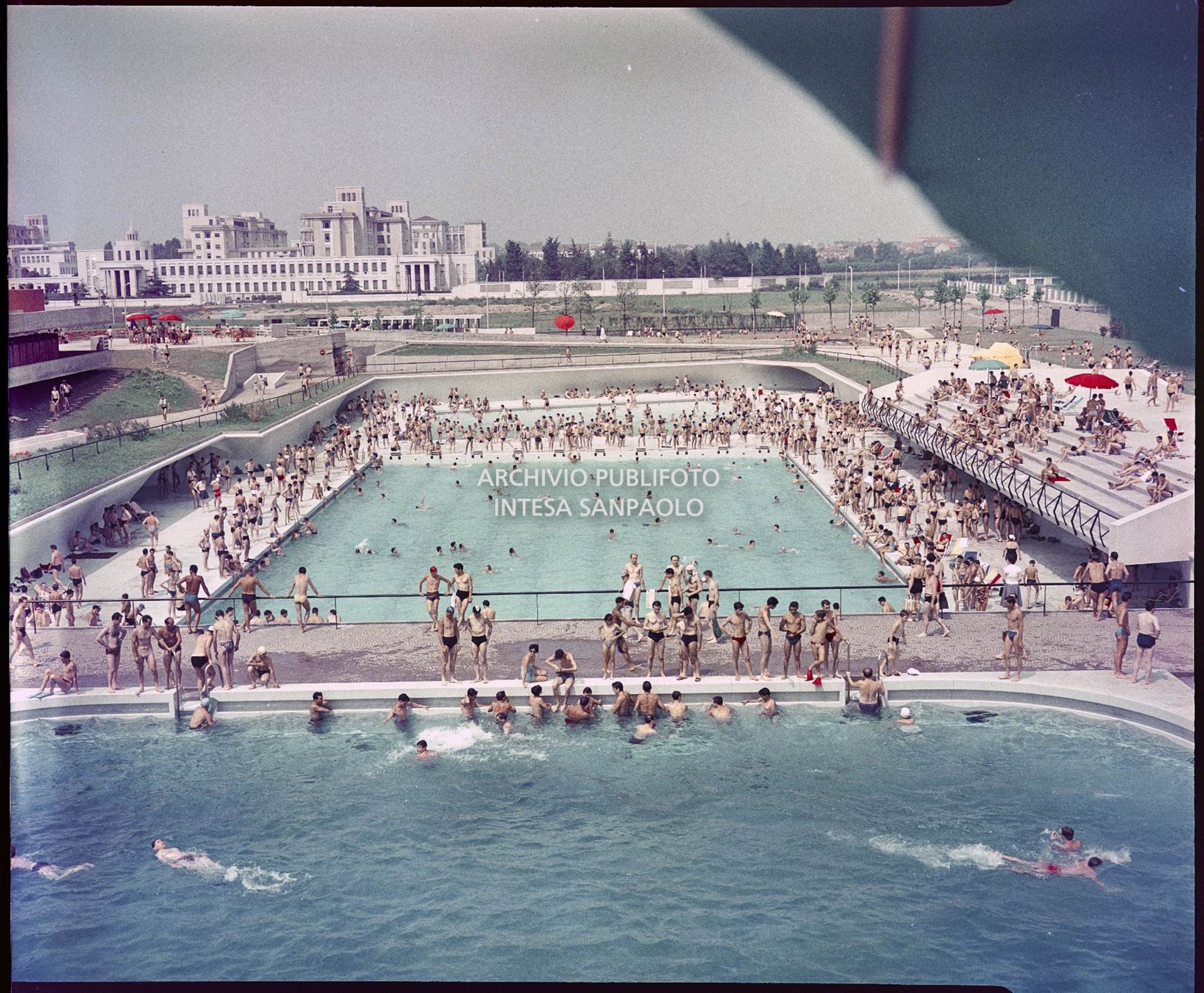 Piscine con bagnanti viste dall'alto. In primo piano è visibile parte di un ombrellone, mentre sullo sfondo l'ospedale Niguarda<br>1590
