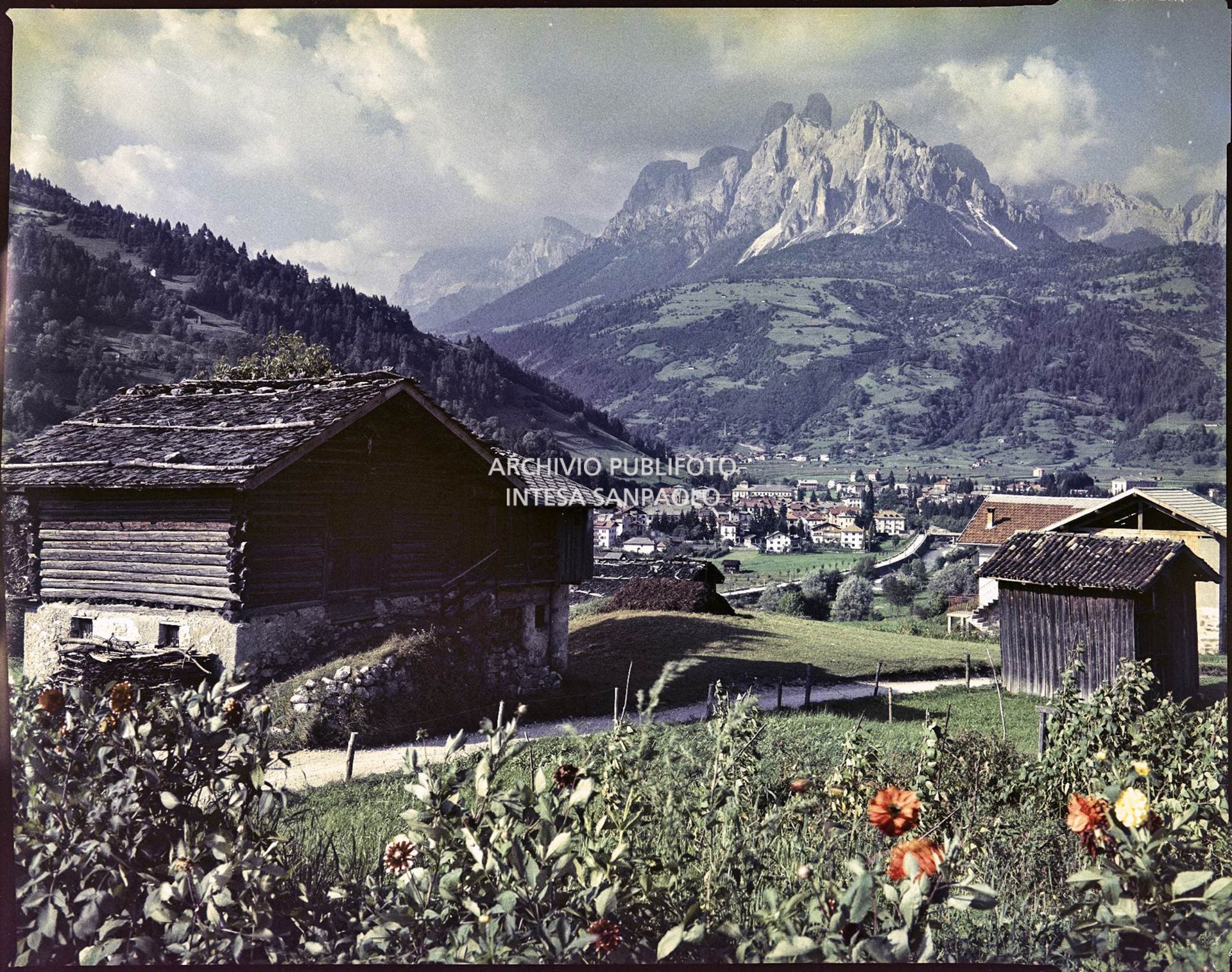 Vista su un paese in una valle. In primo piano una casa tipica in pietra e legno e sullo sfondo le Dolomiti<br>2395