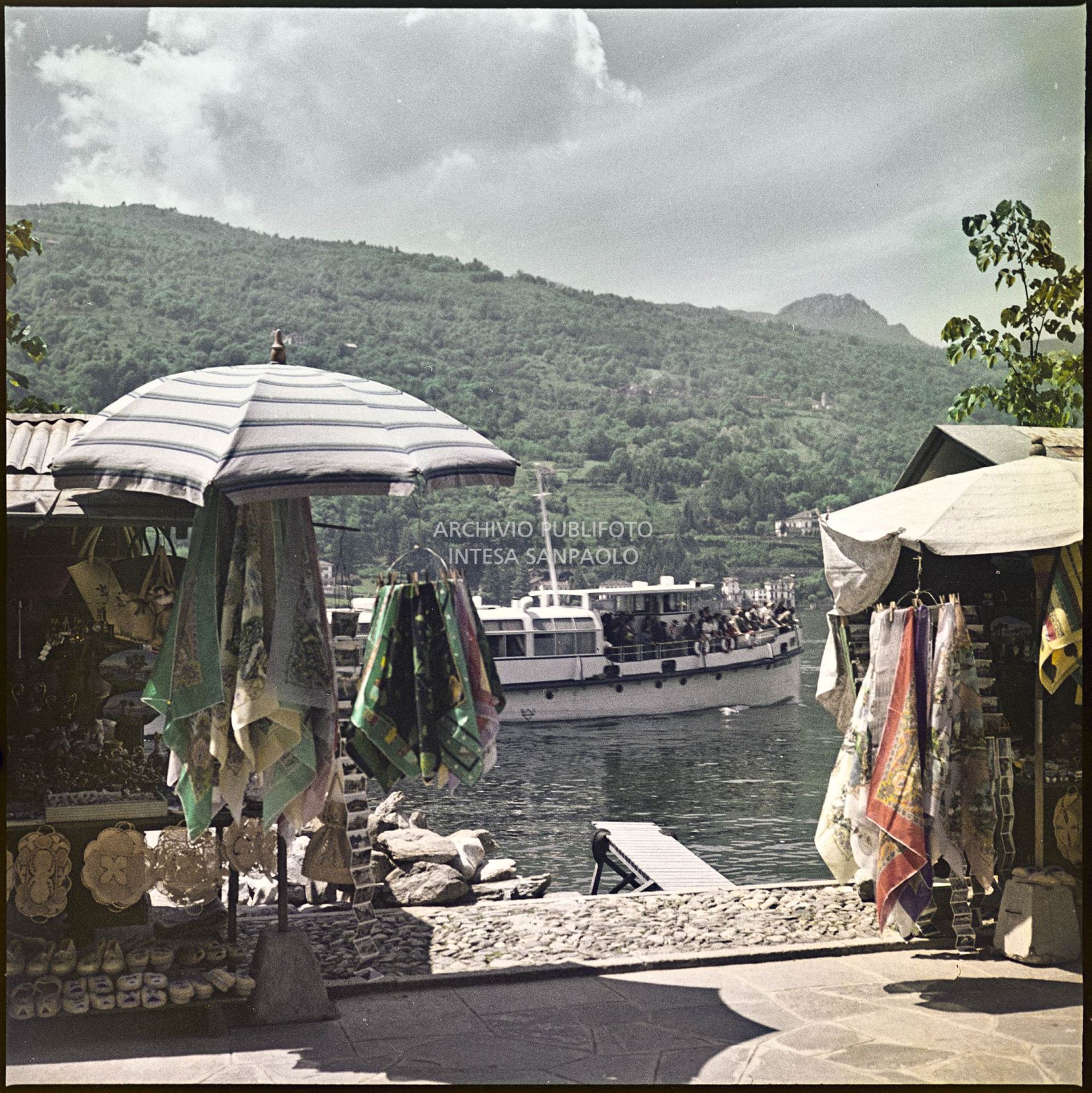 Bancarelle di souvenir sul lungolago. Sullo sfondo un traghetto e le montagne<br>344