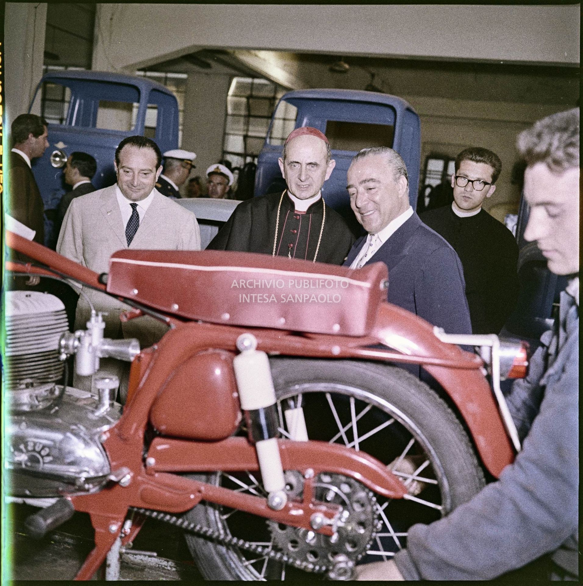 Domenico Agusta ed il cardinale Montini osservano un operaio al lavoro su una motocicletta. Alle loro spalle sono visibili le carrozzerie di alcune vetture ed altre persone, alcune in divisa<br>3458