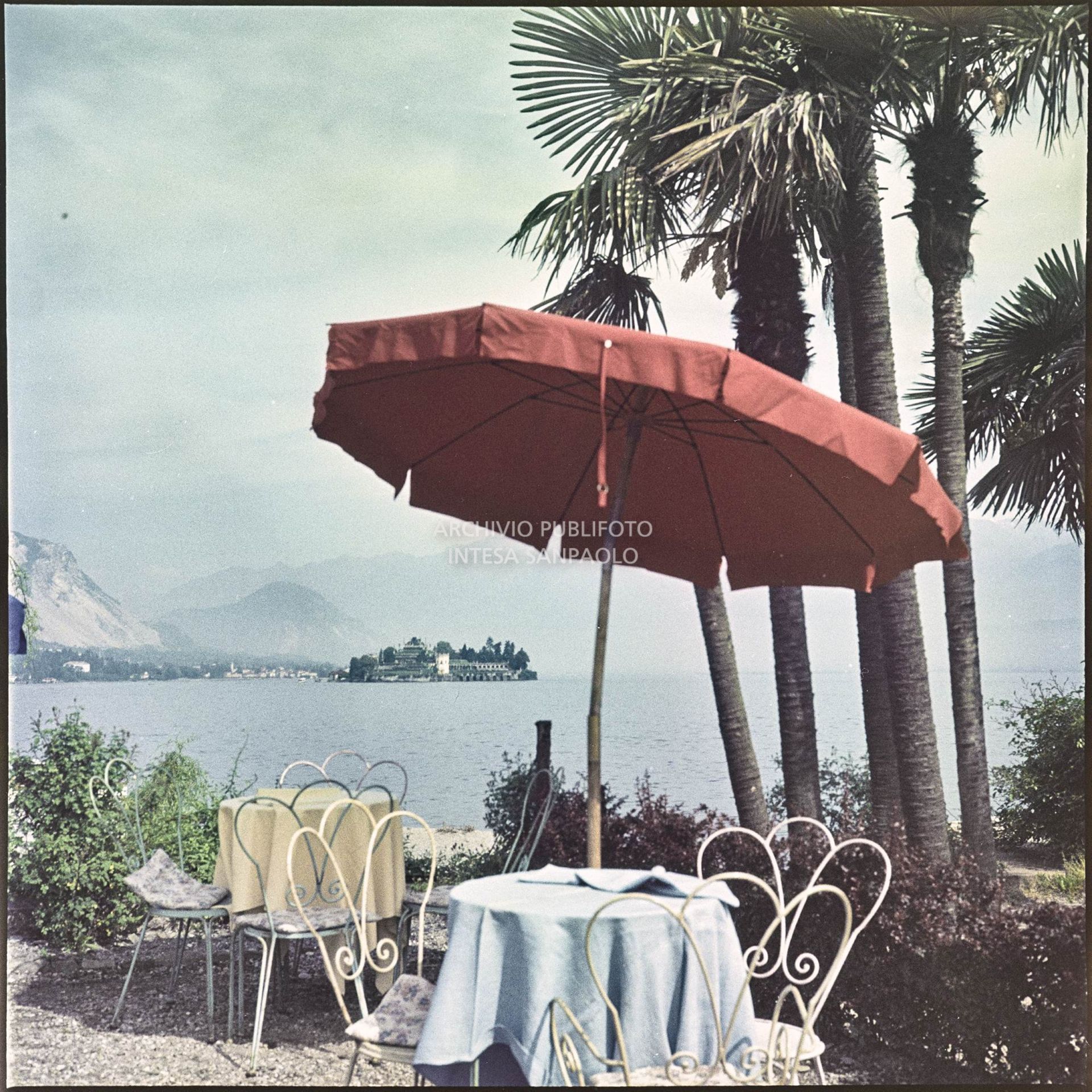 Tavolini ed ombrellone in riva al lago Maggiore. In lontananza è visibile l'Isola Bella<br>350