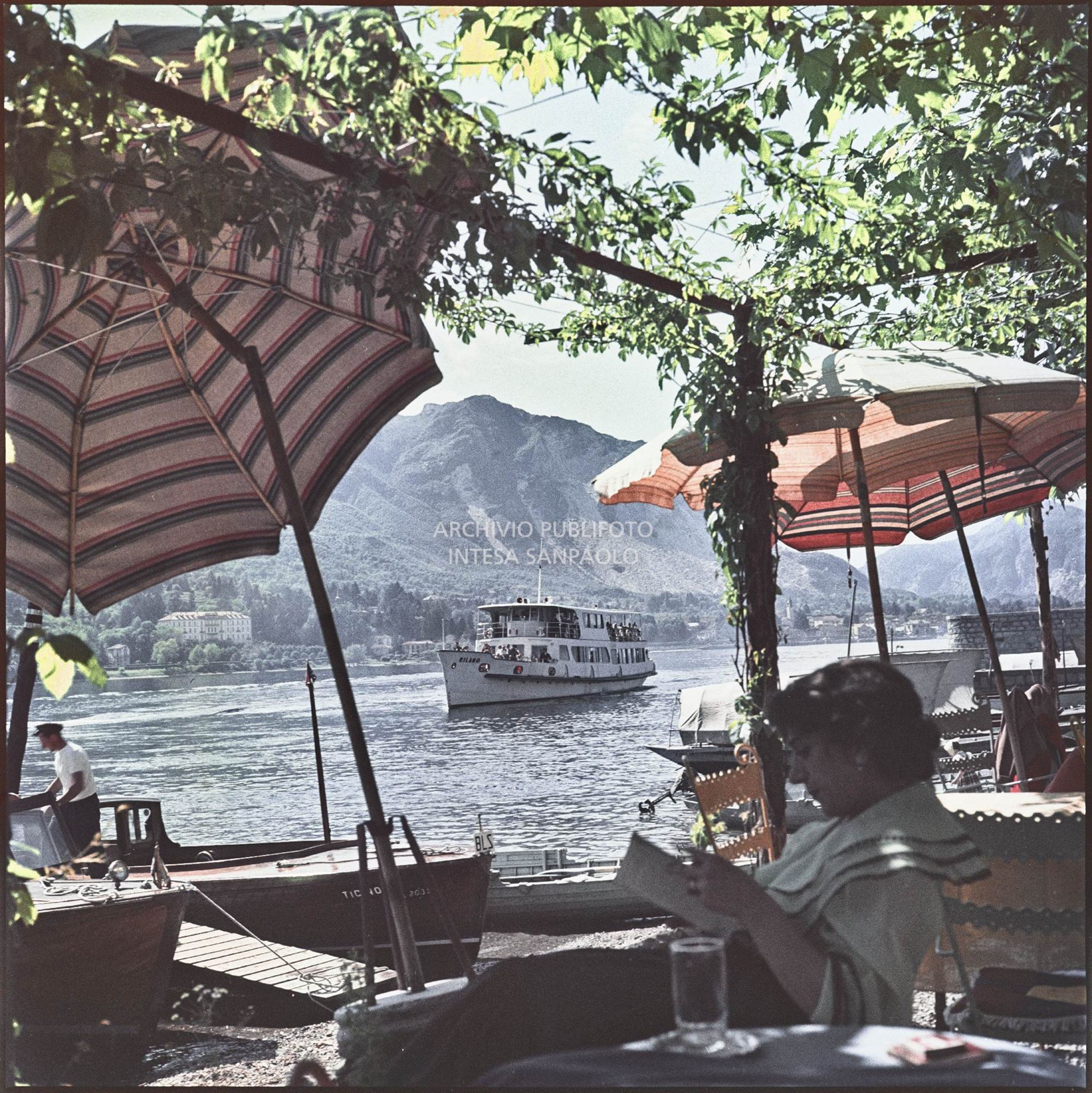 Vista del lago Maggiore e di un traghetto dalla riva dell'Isola dei Pescatori. In primo piano una donna legge un libro sotto ad un pergolato<br>352