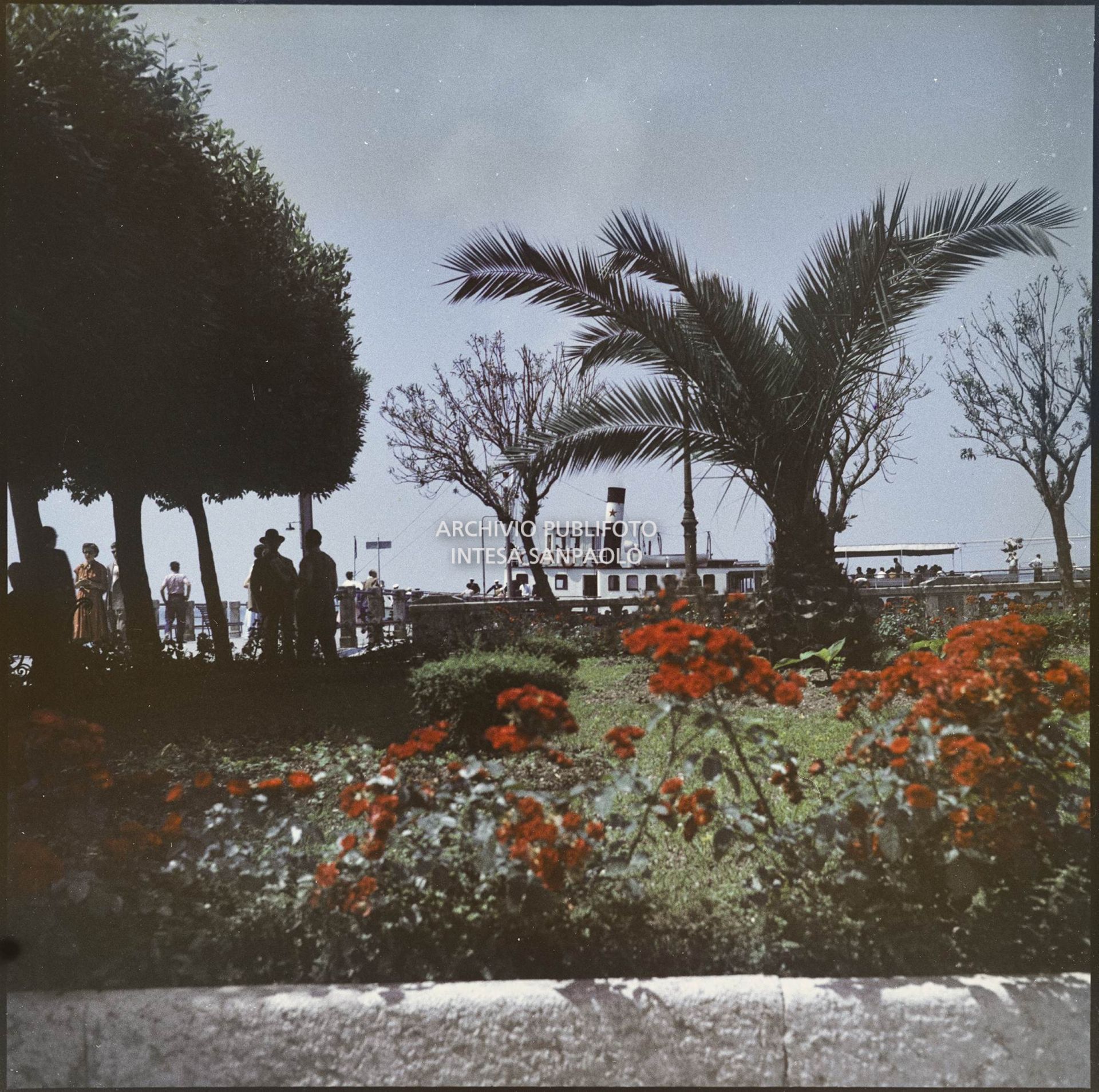 Veduta di un giardino con passanti. In lontananza si intravede un traghetto ormeggiato<br>359