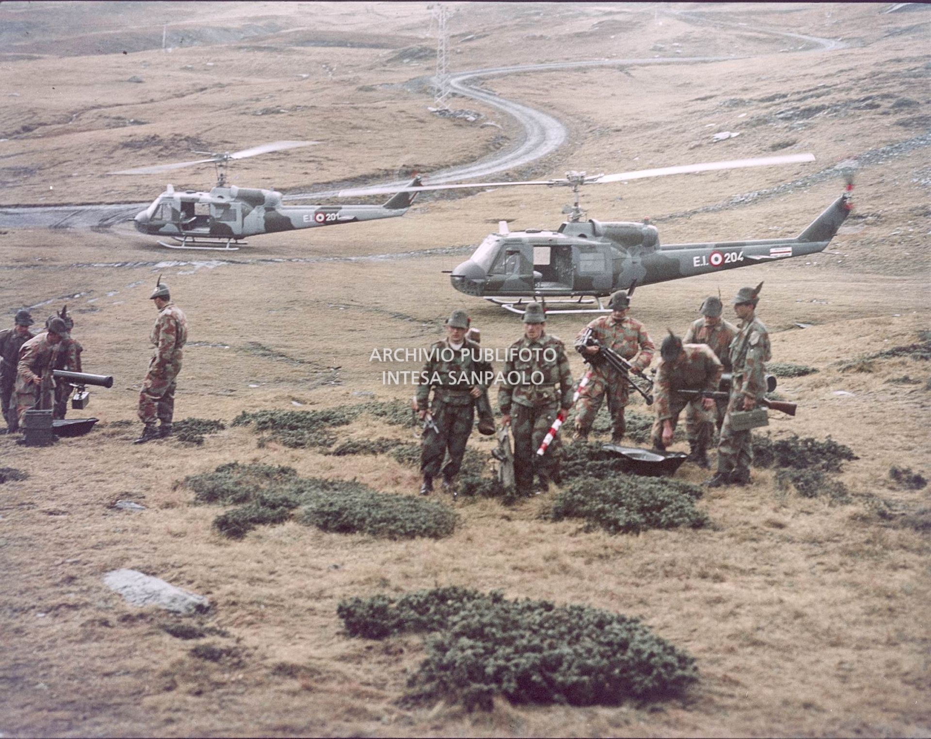Gruppo di Alpini con tuta mimetica, cannoni e fucini. Sullo sfondo due elicotteri militari Agusta-Bell<br>5910