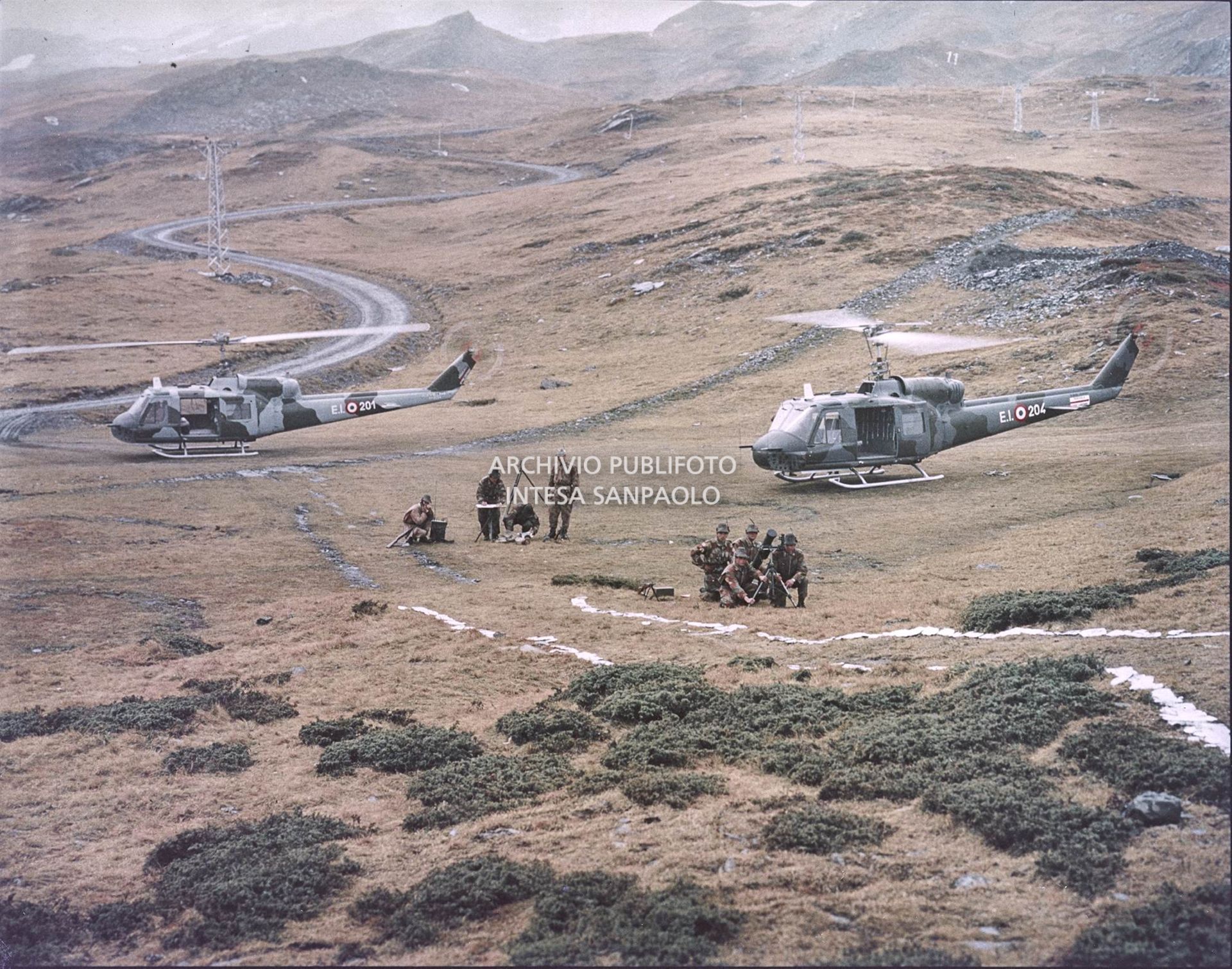 Gruppo di Alpini con tuta mimetica, cannoni e fucini tra le montagne. Sullo sfondo due elicotteri militari Agusta-Bell<br>5919
