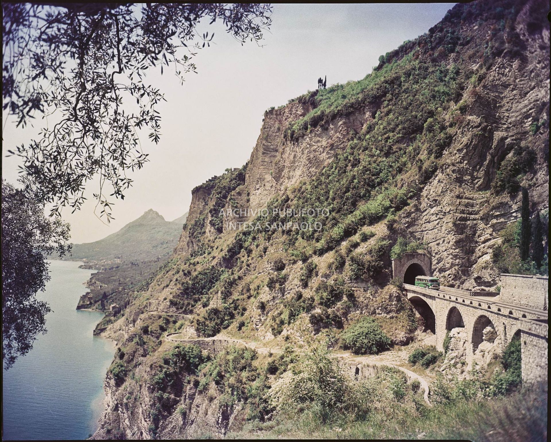 Vista di una strada ed una galleria sul fianco di una montagna in riva al lago. Sulla strada un autobus<br>753