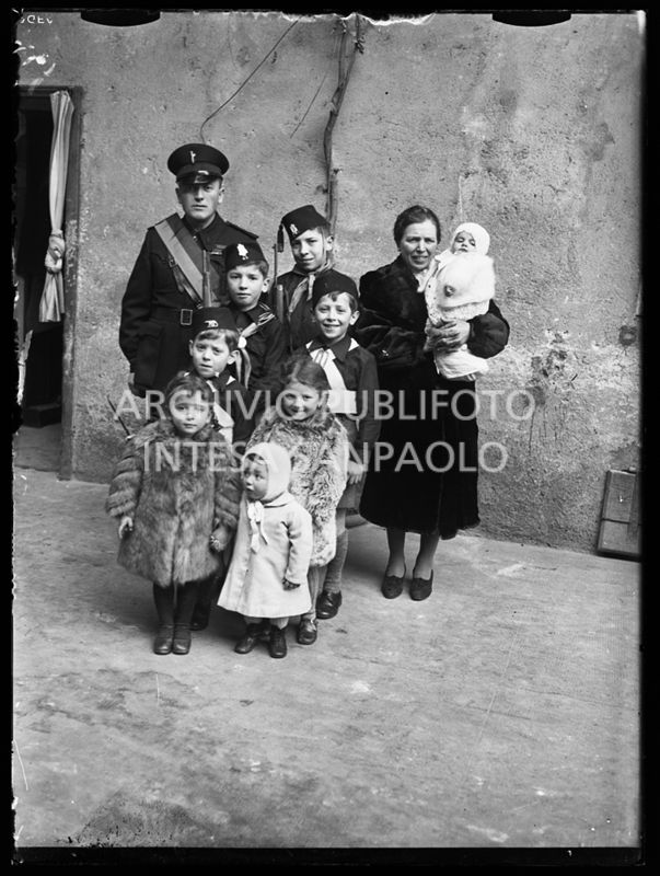 Una coppia, con i suoi otto figli, ritratta davanti all'entrata di casa; i figli maschi indossano divise da balilla e due di loro hanno il moschetto