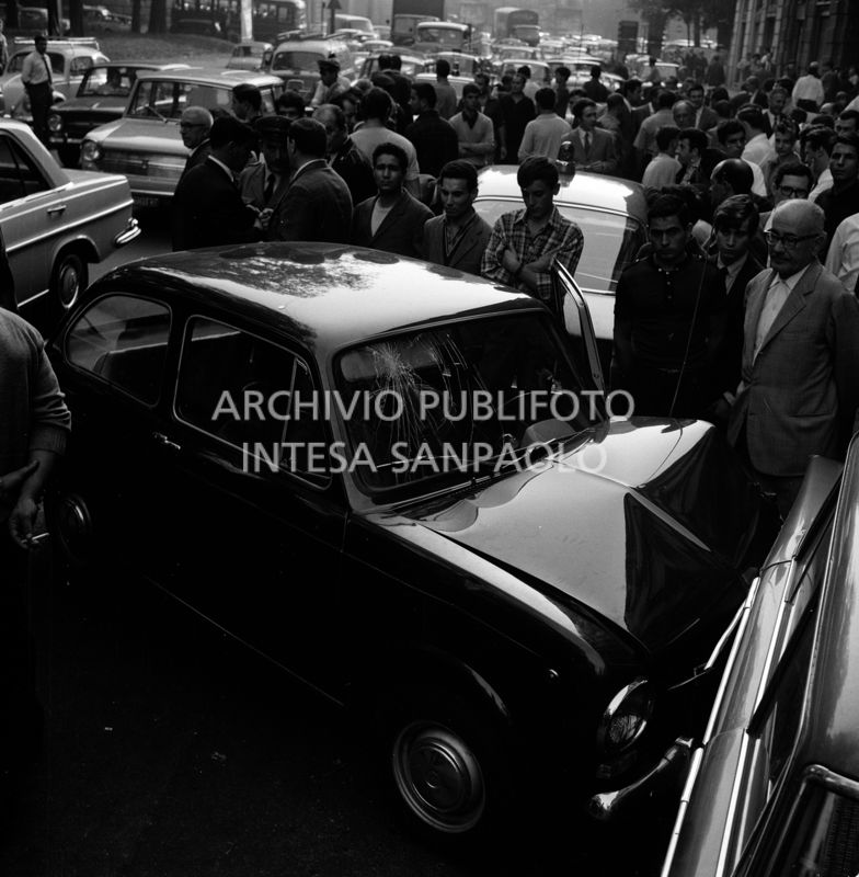 Folla di gente in via Bertani osserva la Fiat 850 che si è scontrata con una Fiat 1500 per evitare l'automobile dei banditi in fuga dopo la rapina all'agenzia del Banco di Napoli di largo Zandonai