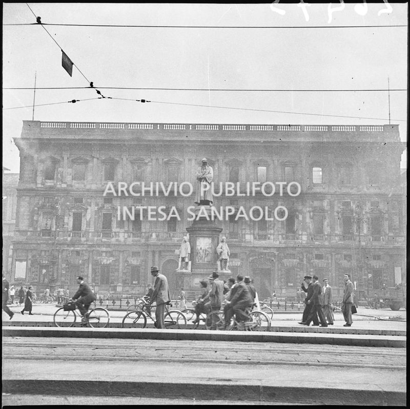 Passanti a piedi e in bicicletta in piazza della Scala a Milano, al cui centro si erge, illeso, il monumento a Leonardo da Vinci. Sullo sfondo Palazzo Marino, a Milano, completamente bruciato e ancora fumante a causa dei bombardamenti, di cui sono rimasti intatti solo i muri perimetrali. Sulla destra, appoggiata al palazzo, la scala dei vigili del fuoco