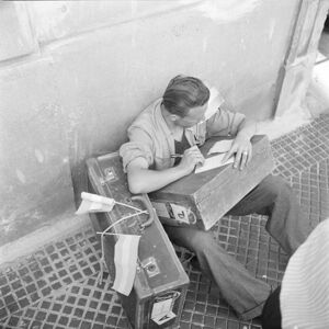Un emigrante italiano a Genova, seduto per terra a fianco alle valigie, scrive su un foglio in attesa di partire per l'Argentina