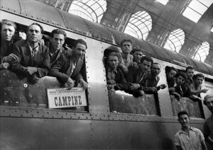 Emigranti, affacciati al vagone ferroviario di un treno, in partenza per le miniere del Belgio, dalla Stazione Centrale di Milano. Sul treno il cartello: "Federation des charbonnages 66, rue des Colonies, Bruxelles. Campine"