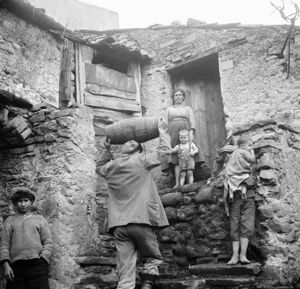 Africo, un ragazzo sale le scale di una casa trasportando una botte sulla testa; intorno a lui dei bambini e una donna