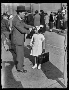 Un uomo sistema i capelli a una bambina, col grembiule e la cartella, davanti a scuola