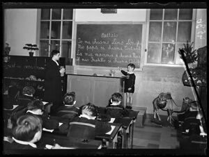 Bambini, col grembiule, in classe durante una lezione: uno di loro è interrogato dall'insegnante alla lavagna