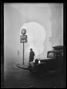 Scorcio di un angolo di Milano immerso nella nebbia: sono riconoscibili un uomo, sotto un orologio pubblico, e un'automobile con i fari accesi