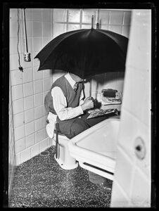 Un uomo, seduto sul water, legge il giornale riparandosi sotto un ombrello aperto