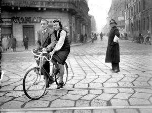 Un uomo e una donna a bordo di un particolare modello di bicicletta in una via di Milano, poco più in là un vigile urbano li guarda incuriosito