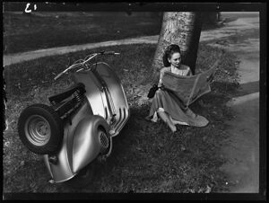Una donna legge il quotidiano appoggiata a un albero di un parco con accanto una Vespa 125 V1T