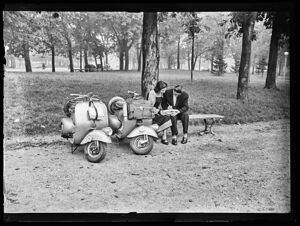Una coppia di ragazzi, seduti su una panchina in un parco di Milano, studia. Accanto sono parcheggiate due Vespe Bacchetta 125