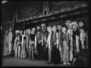 Un burattinaio, affacciato al sipario del teatrino, posa circondato dalla sua compagnia di marionette