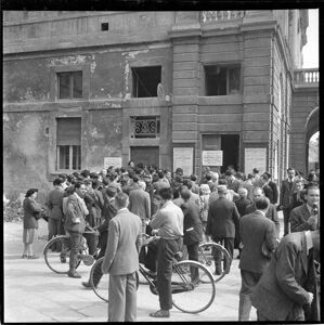 Persone stazionano davanti a una delle entrate laterali del Teatro alla Scala sul quale sono affissi il manifesto del concerto di Arturo Toscanini per la riapertura del Teatro, ricostruito dopo la guerra, e alcuni cartelli che indicano le modalità di acquisto dei biglietti