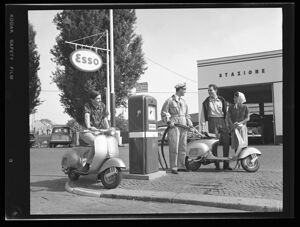 Distributore Esso, benzinaio fa il pieno ad una Vespa con accanto una coppia;  in attesa un altro cliente in Vespa