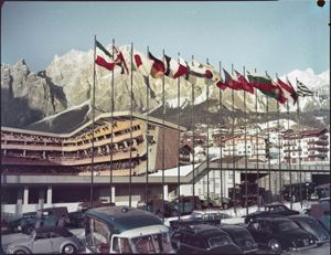 In primo piano delle automobili parcheggiate, tra cui in furgone con il marchio Martini; sullo sfondo le Dolomiti<br>